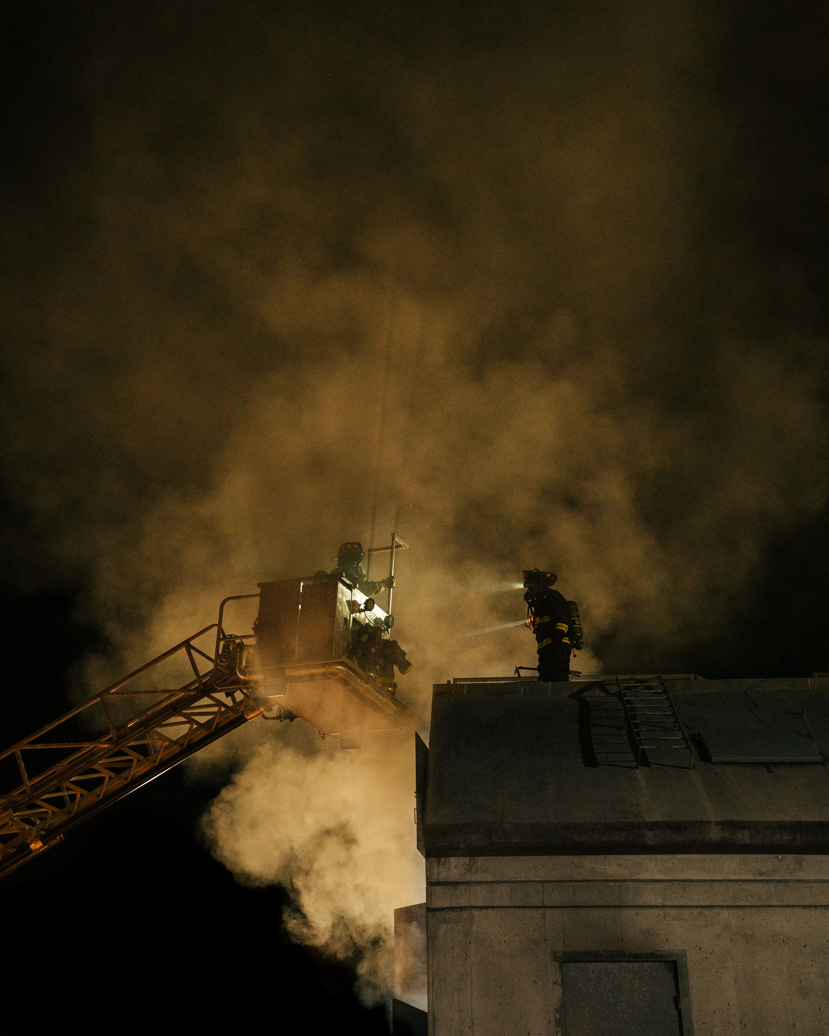 Firefighters working a rooftop through heavy smoke at night, one on the ladder bucket, one on the roof